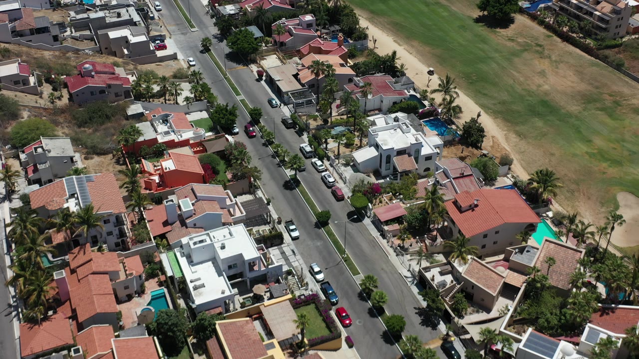 Residential aerial with street, cars, rooftops, pools, and an open field.