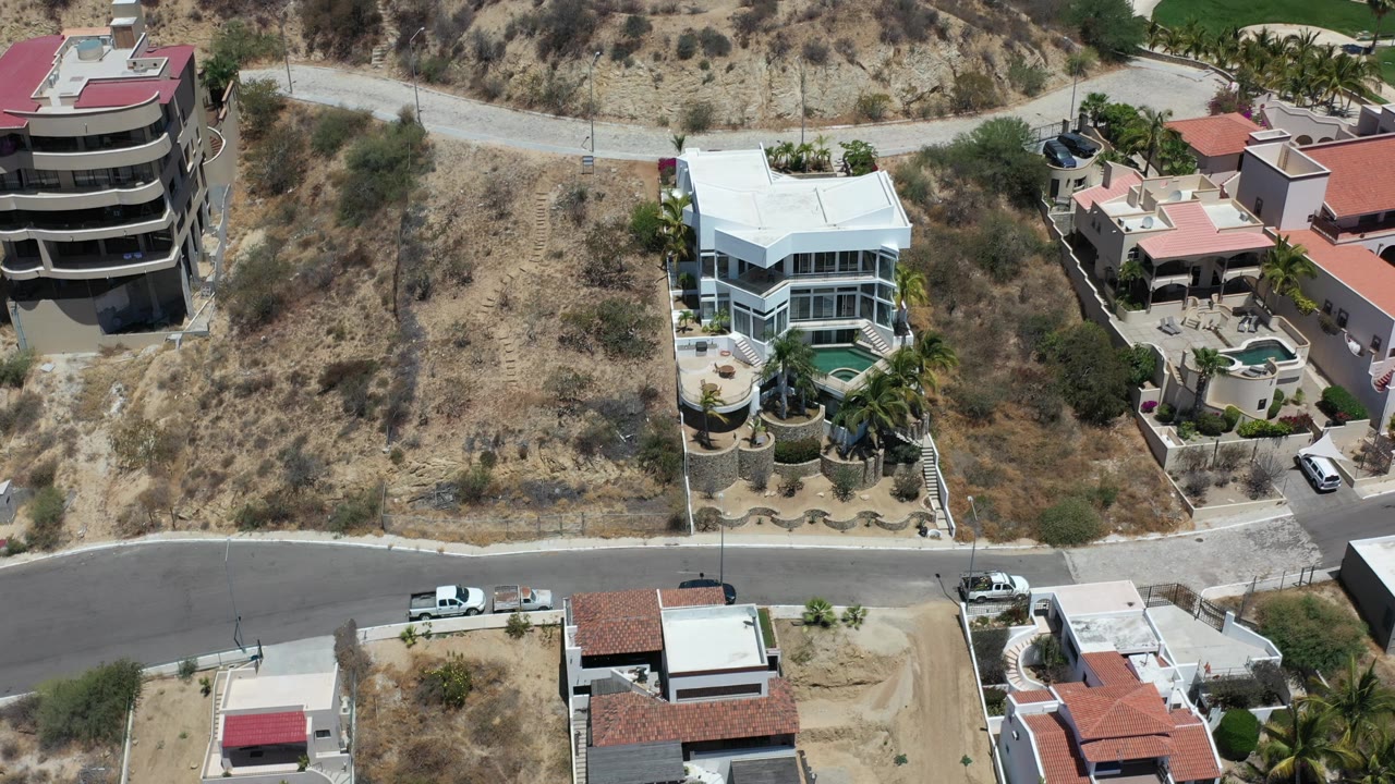 Aerial Los Cabos residential view with dry open lots in the center, a paved street across the frame, white and terracotta-roof homes, parked vehicles, and no visible people.