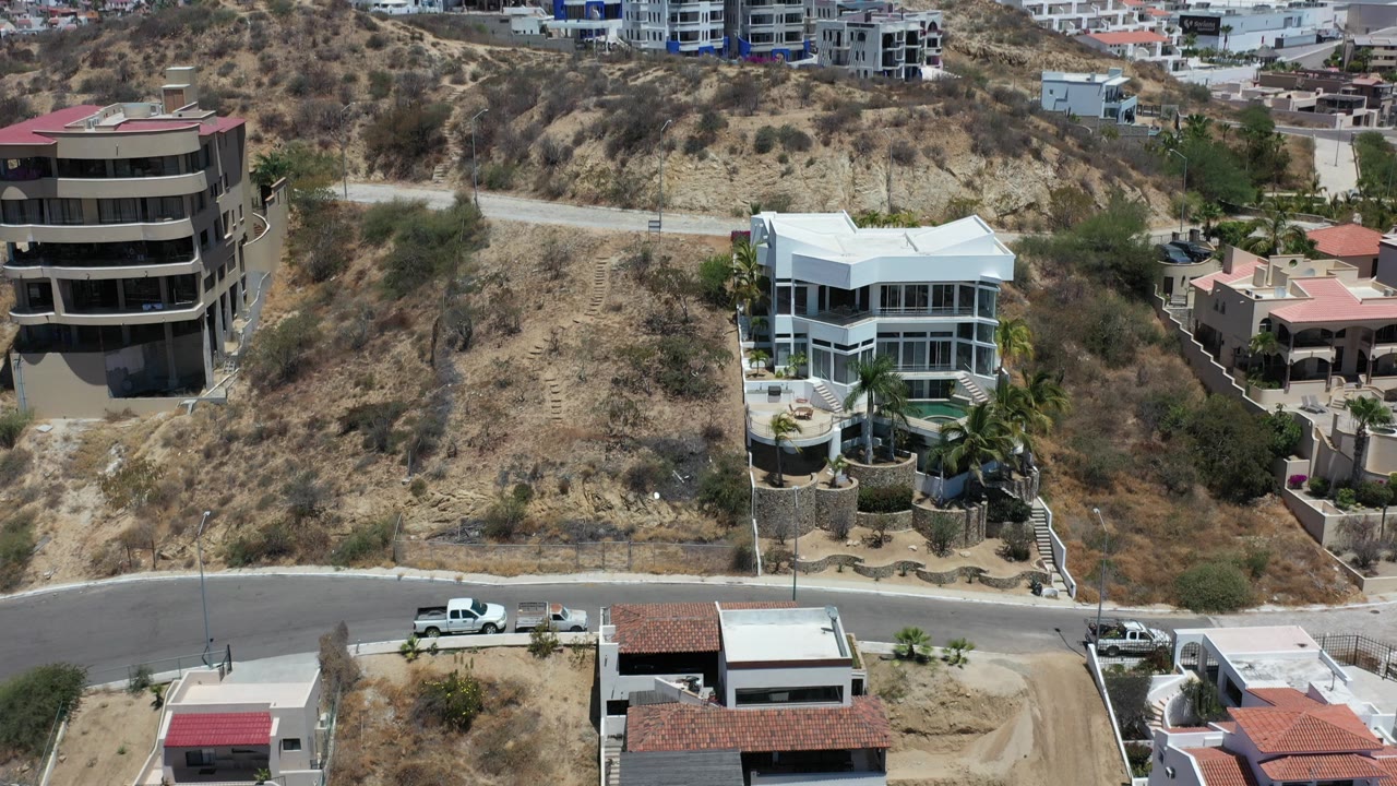 Aerial view in Los Cabos, Mexico, showing a white multi-story mansion on a dry hillside beside other homes, a road, parked vehicles, and no visible people.