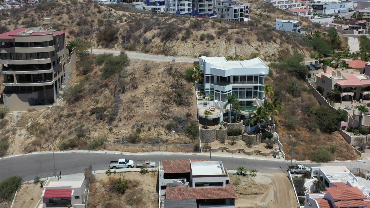 Aerial frame in Los Cabos, Mexico, centered on a white hillside mansion with a dirt slope, adjacent multi-story homes, foreground roofs, and no visible people.