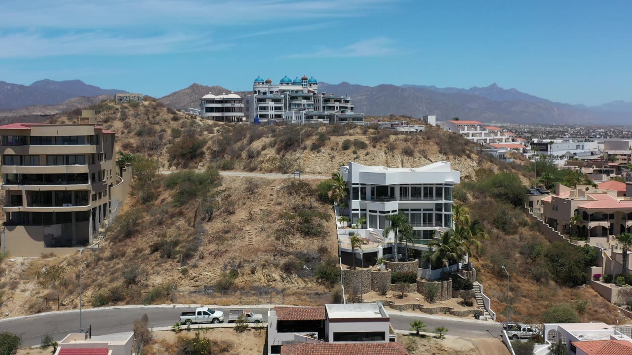 Aerial view slightly farther along the hillside, showing white and tan modern homes, scrub-covered slopes, a road at the base, and no visible people in Los Cabos, Mexico.