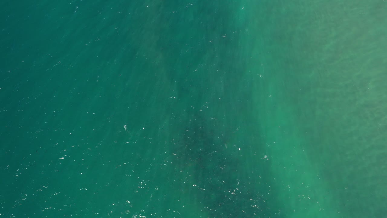 Opening overhead view of blue-green ocean water in Los Cabos, Mexico, with pale submerged streaks near the upper right and no visible people.