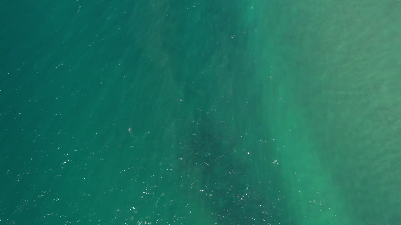 Overhead Los Cabos ocean frame with darker green bands across the water, scattered surface sparkle, and no visible people.