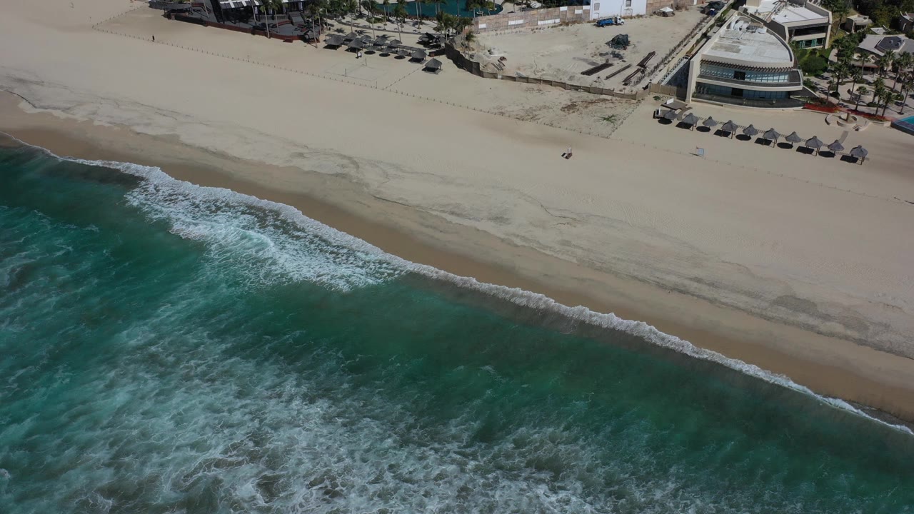 A high aerial view in Los Cabos, Mexico shows turquoise surf rolling onto a wide pale beach, with beachfront buildings and umbrellas near the top of frame and tiny distant people on the sand.