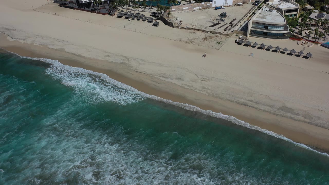 The Los Cabos shoreline fills the frame with darker turquoise water below, a thin white wave line at mid-frame, open sand, umbrellas, and beachfront buildings along the upper edge.