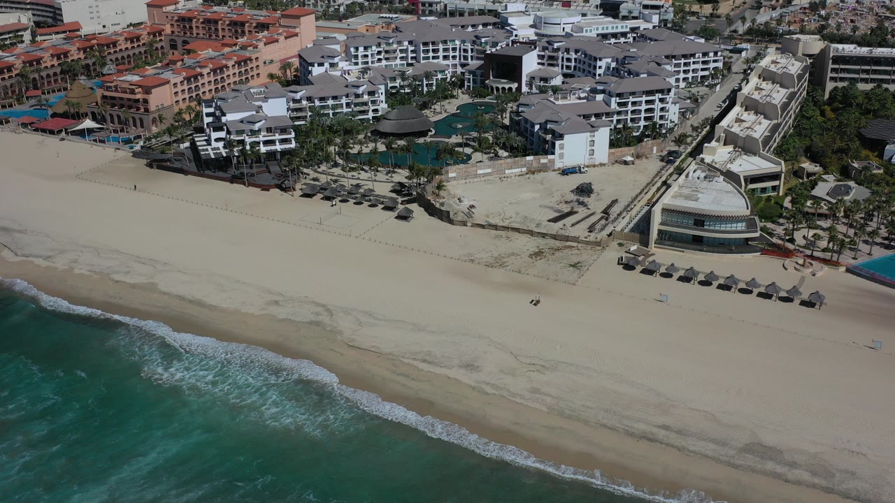 Aerial view of surf, white sand beach, umbrellas, and beachfront buildings.