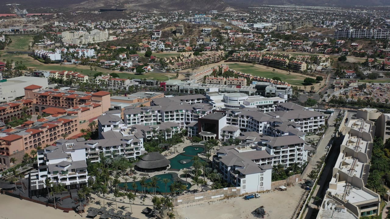 High aerial frame over a Los Cabos resort, showing gray and white hotel buildings, palm landscaping, turquoise pools, surrounding roads, and dry hills in the distance with no visible people.