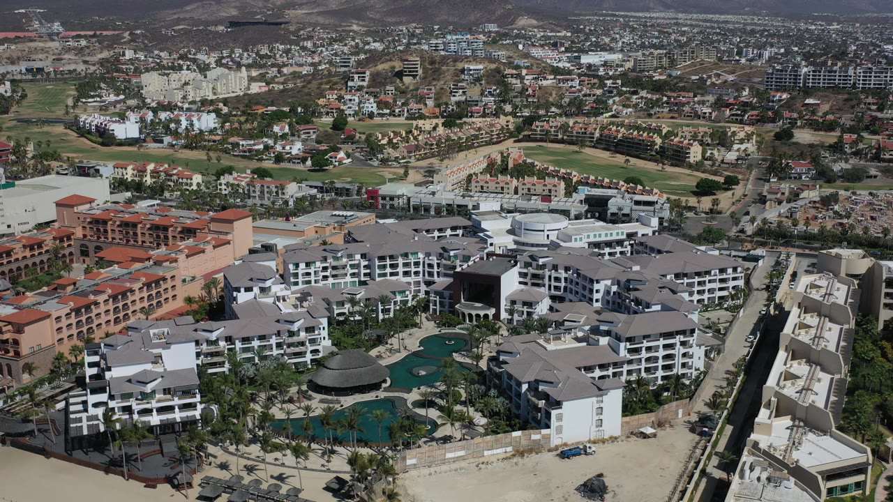 Aerial view in Los Cabos with the resort centered lower in frame, turquoise pools and palms below, orange hotel buildings to the left, roads to the right, and no visible people.