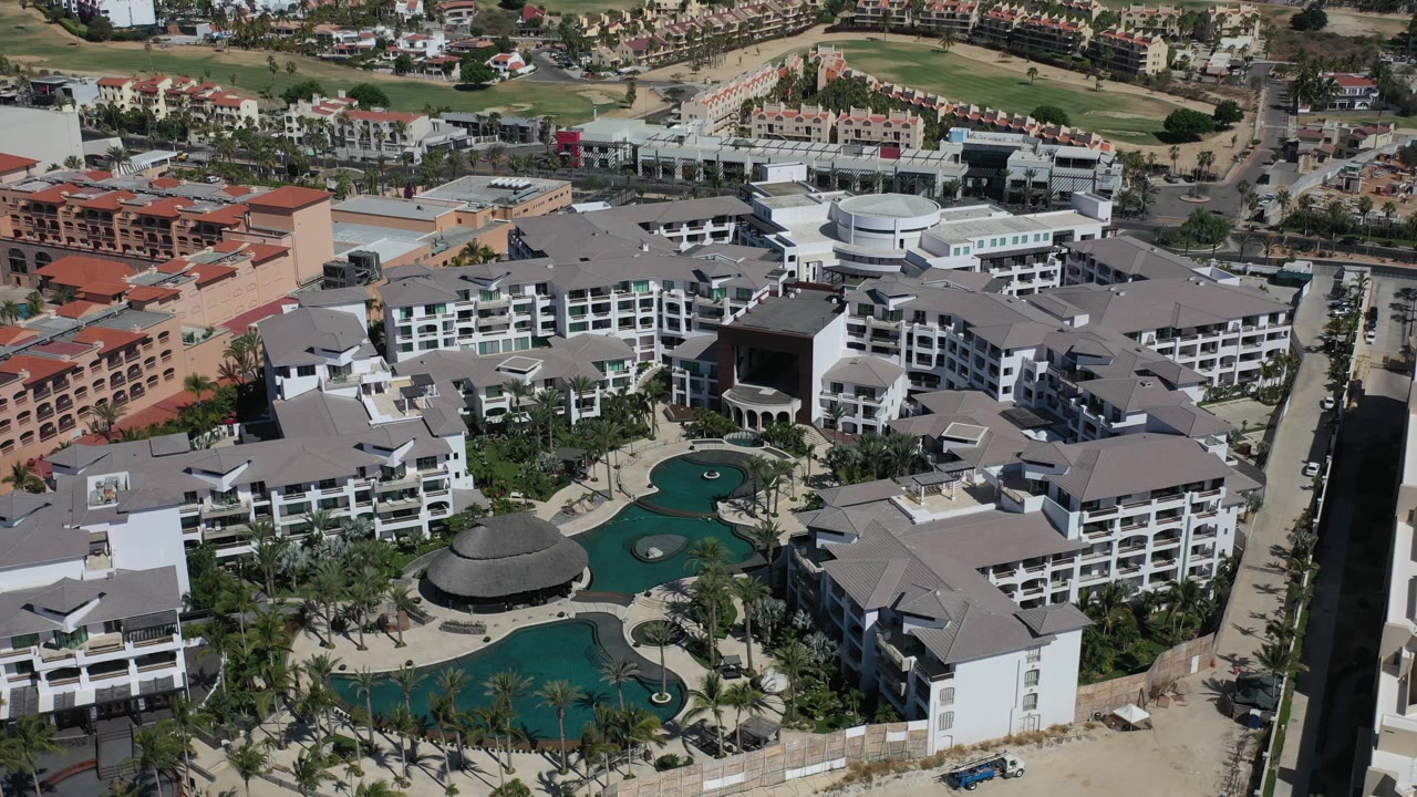 Overhead Los Cabos frame with white multi-story condos, gray roofs, a rounded dark-roof pavilion, turquoise pools, palms, and no visible people.