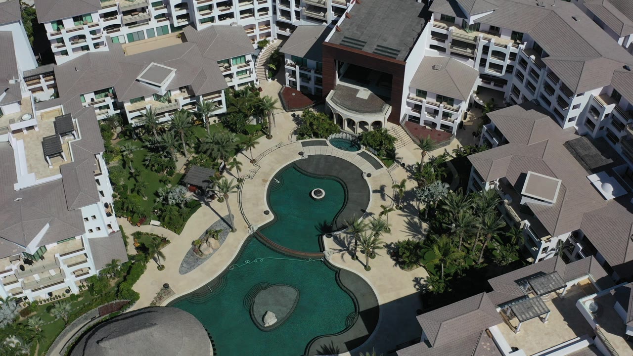 Downward aerial frame over a Los Cabos hotel resort courtyard, showing curved green pools, palm landscaping, pale paving, white balconies, gray roofs, and no visible people.