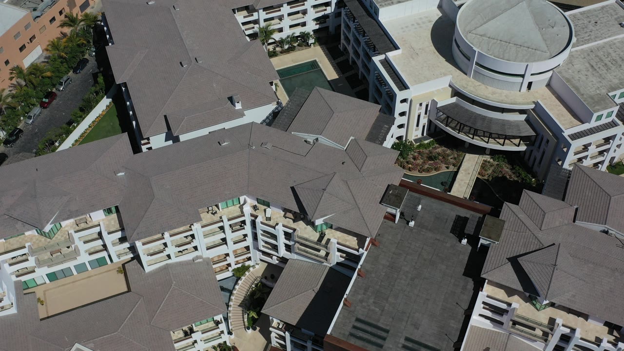 Overhead aerial frame in Los Cabos, Mexico showing gray rooftops, white balcony rows, courtyards, palms, and a circular building, with no visible people.