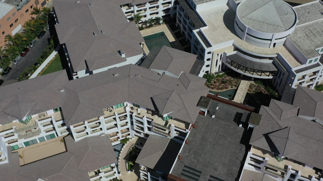 Second aerial frame over Los Cabos, Mexico with the gray roof mass centered, white apartment-style balconies below, and the round building near the upper right, with no visible people.