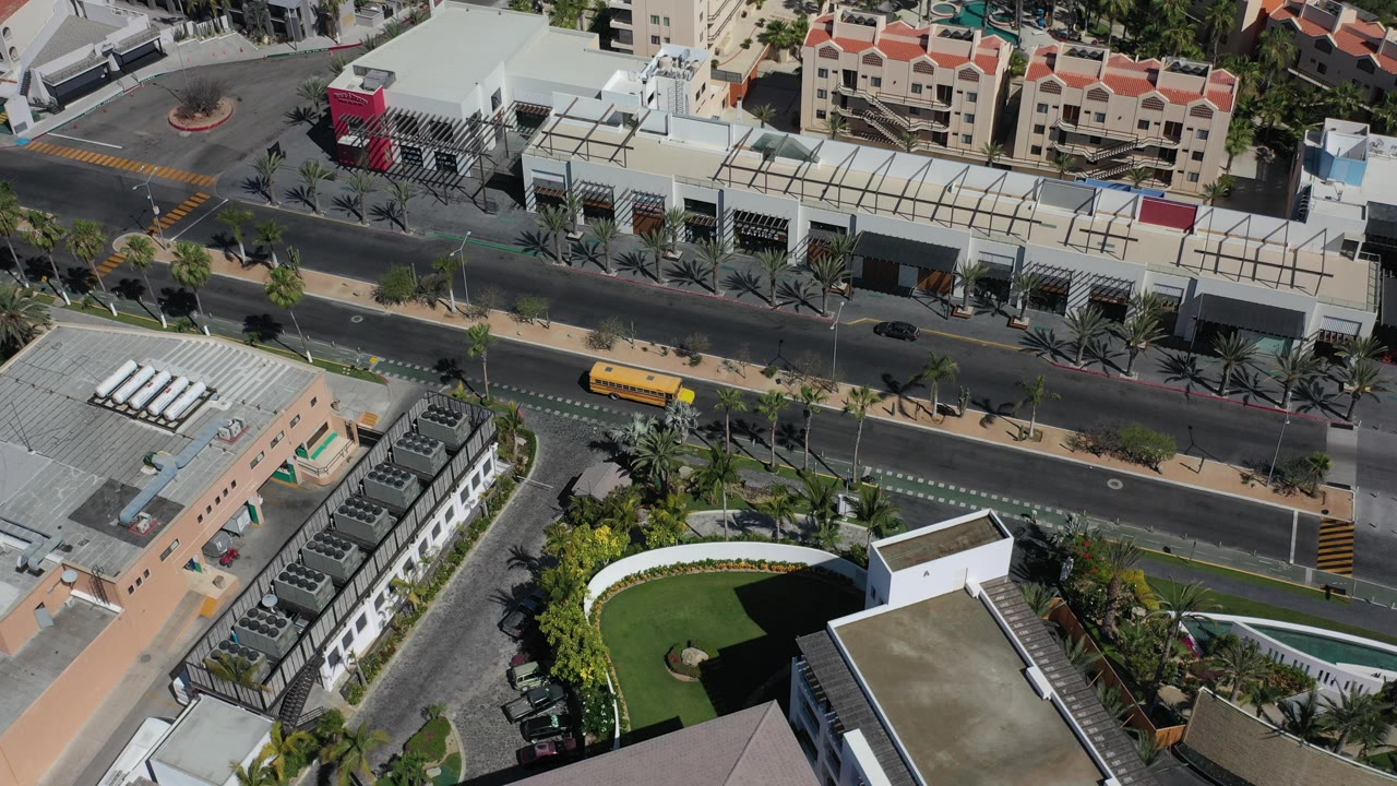 Aerial view of a Los Cabos road, condo buildings, palms, and a yellow bus.