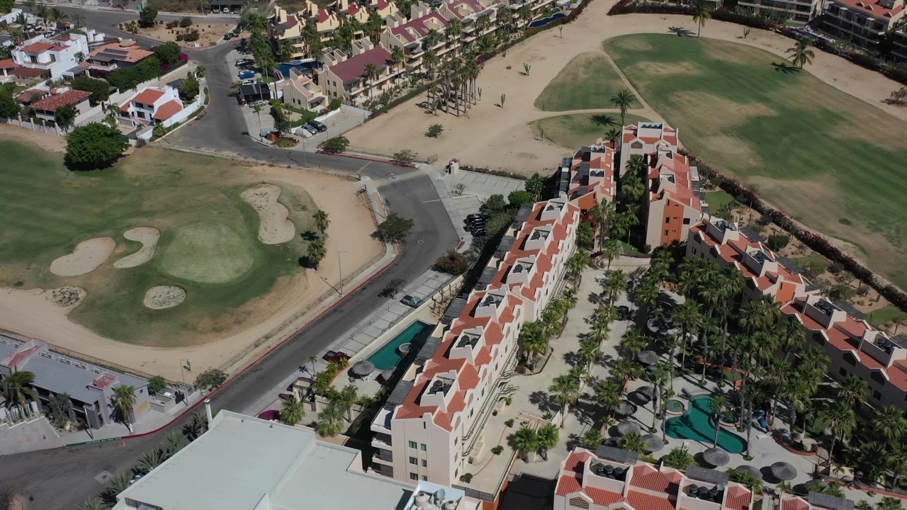 Overhead Los Cabos frame with a large green fairway, pale sand bunkers, palm-lined housing, nearby roads, and no visible people.
