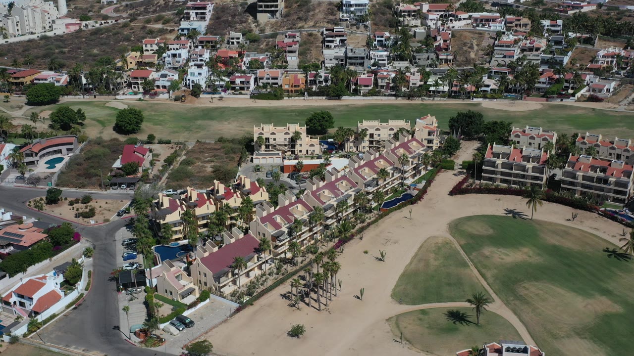 A high aerial frame over Los Cabos shows red-tile-roof homes, a curved street, tan dry ground, green fairway sections, hillside housing in the distance, and no visible people.