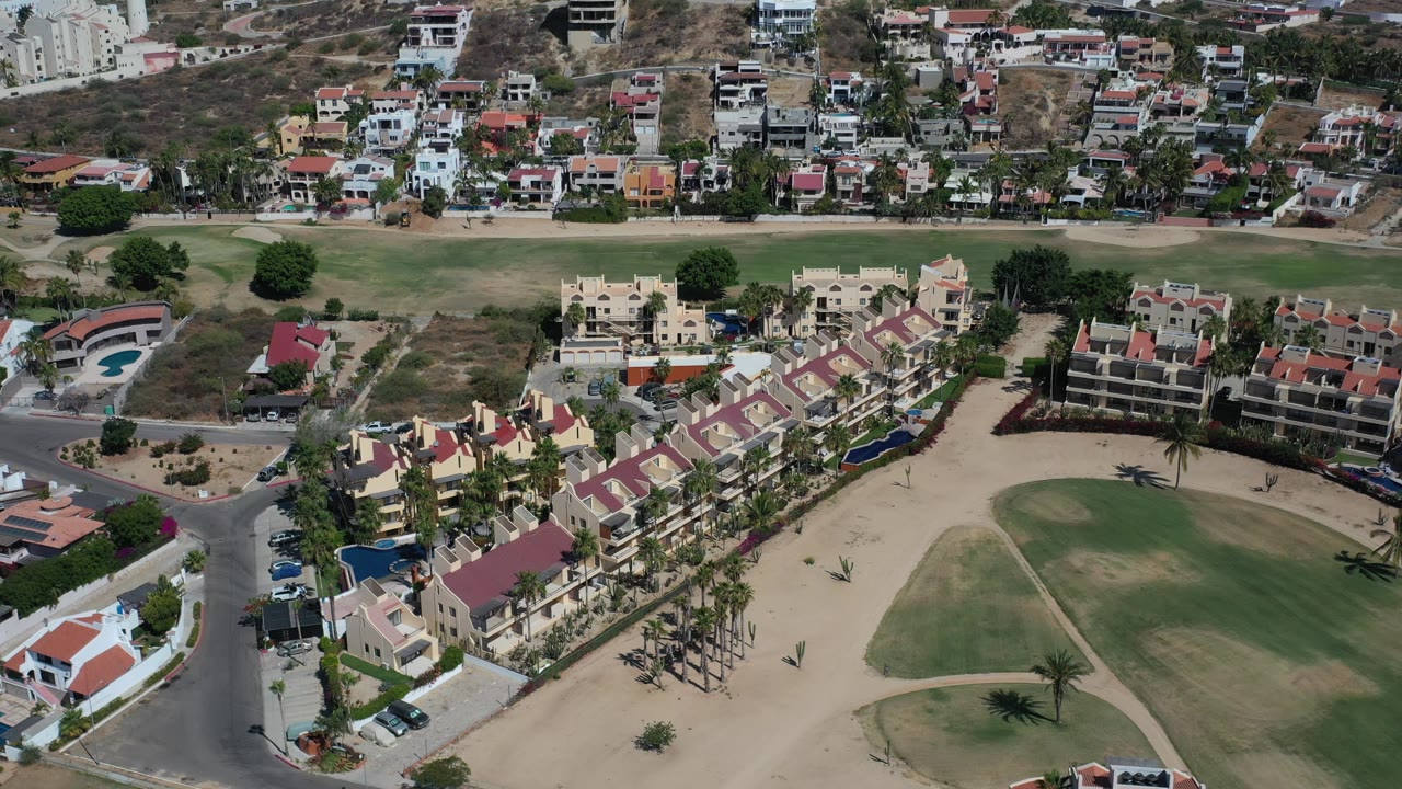 The second frame holds a high view across Los Cabos residences, with a row of red-roof stucco buildings below, palms along the street, scrubby hillside development above, and no visible people.