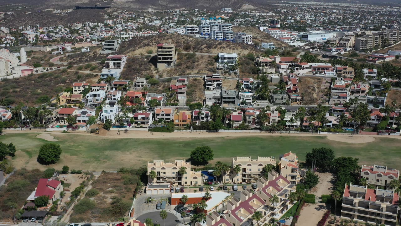 Aerial Los Cabos frame with the grassy foreground stretching across the lower image, compact houses and roadside trees in the middle, and hillside buildings in the distance, with no visible people.