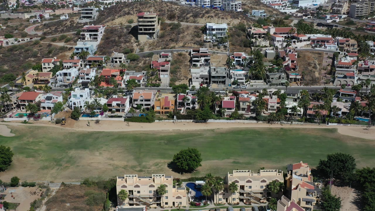 High aerial frame over Los Cabos, Mexico, showing a strip of beachfront homes, a road, dry hillside development, green open space, sandy beach, and shallow water with no visible people.