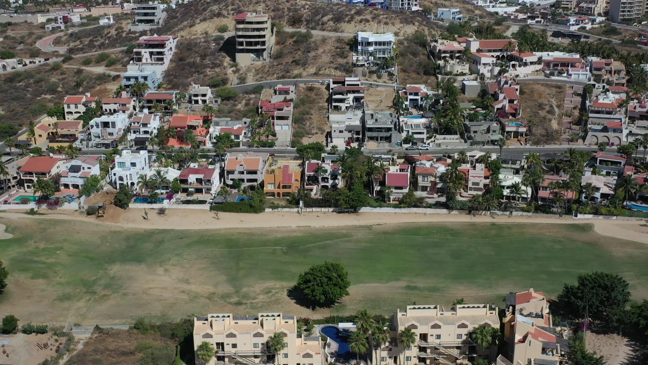 Aerial coastal neighborhood with homes, palms, road, hillside, grass, and water.