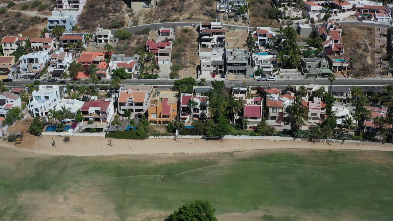 Aerial view in Los Cabos, Mexico with beachfront homes, palms, a horizontal road, sandy beach, shallow water, and no visible people.