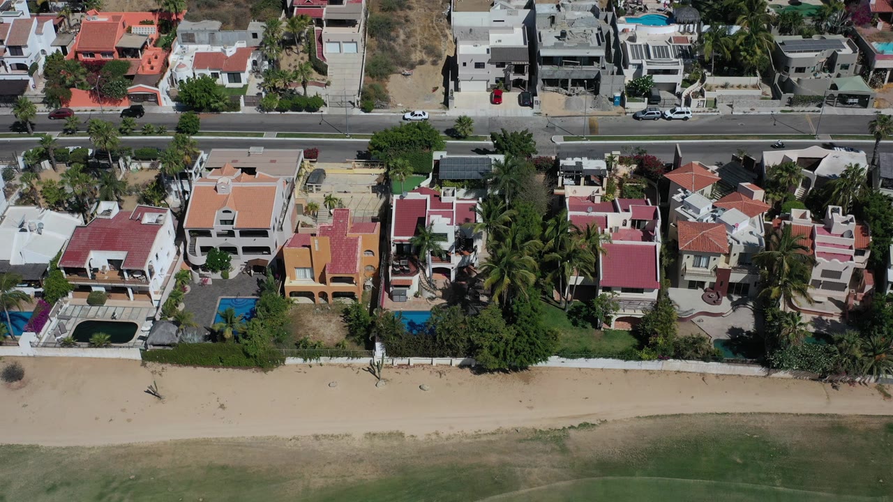 Overhead aerial frame in Los Cabos showing beachfront private homes, a road at the top, blue pools, palms, a sandy strip, and no visible people.