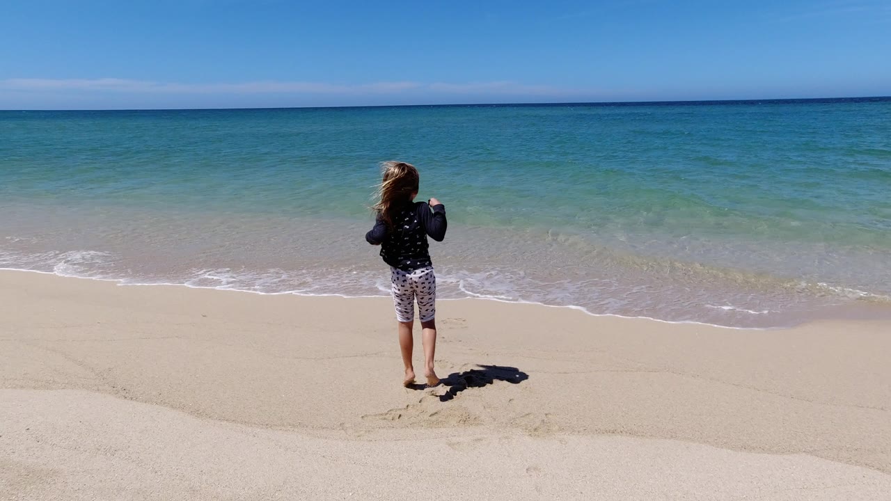 Child standing on sandy beach facing ocean waves.