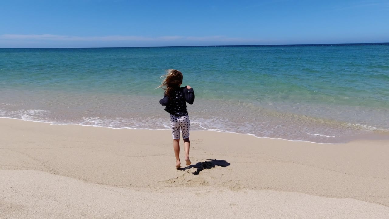 The child remains near the center of the wide frame, turned toward the turquoise ocean as a thin line of foam reaches the shore in Los Cabos.