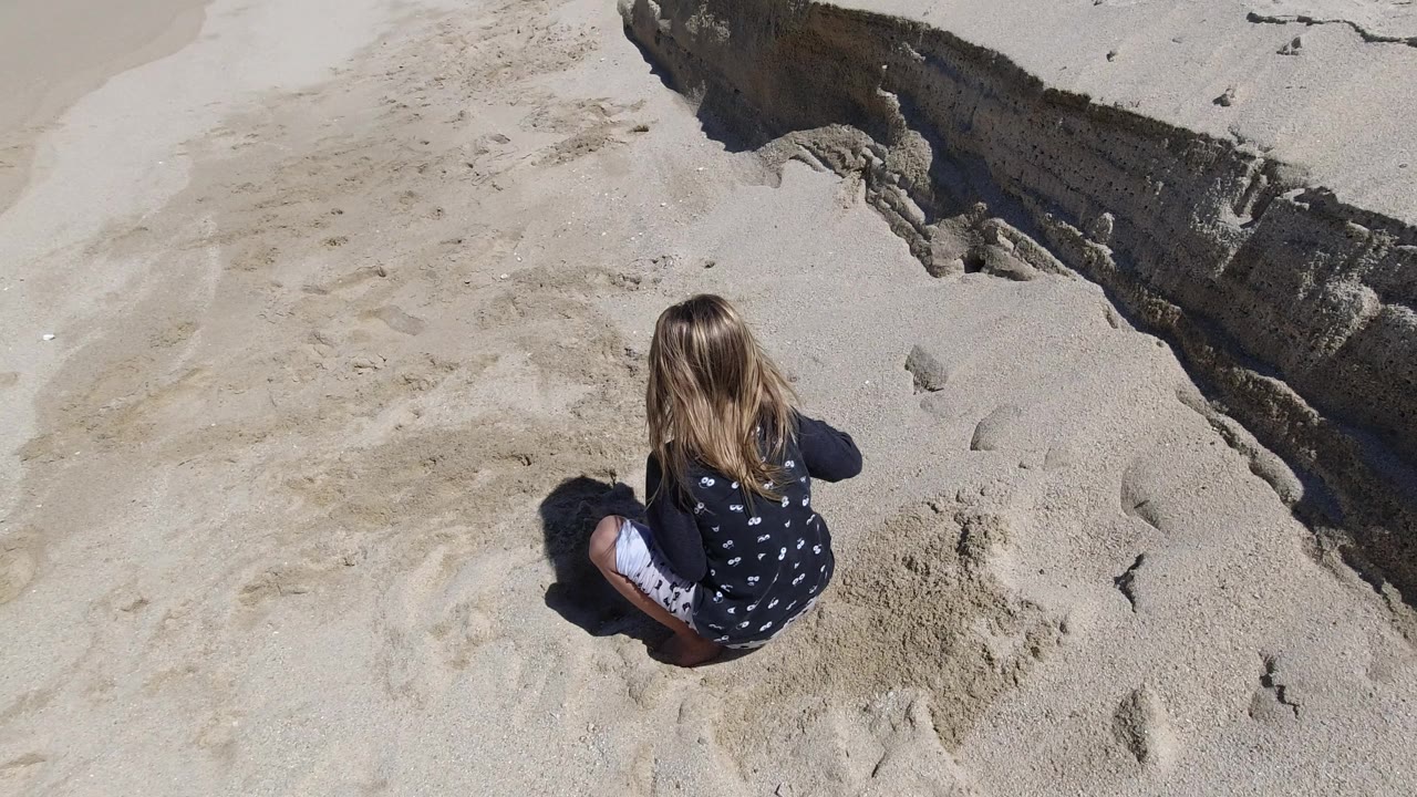 Overhead frame of a young girl seated with her back to camera on pale sand in Los Cabos, Mexico, near a dark eroded sand edge at the upper right.