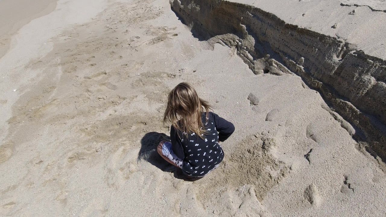Girl sitting alone on sand beside a raised beach edge.