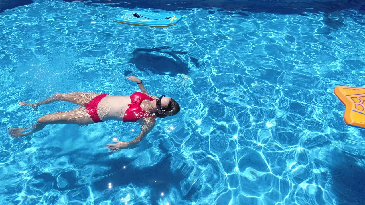 Adult woman in a red swimsuit and sunglasses floats on her back in a bright blue pool in Los Cabos, Mexico, with a blue inflatable near the top edge.