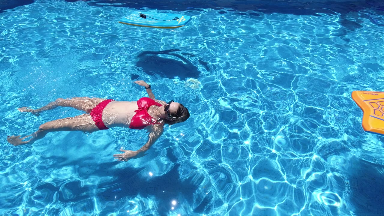 Adult woman continues floating on her back in the Los Cabos pool, with her arms loose at her sides and a bright orange float cropped along the right edge.