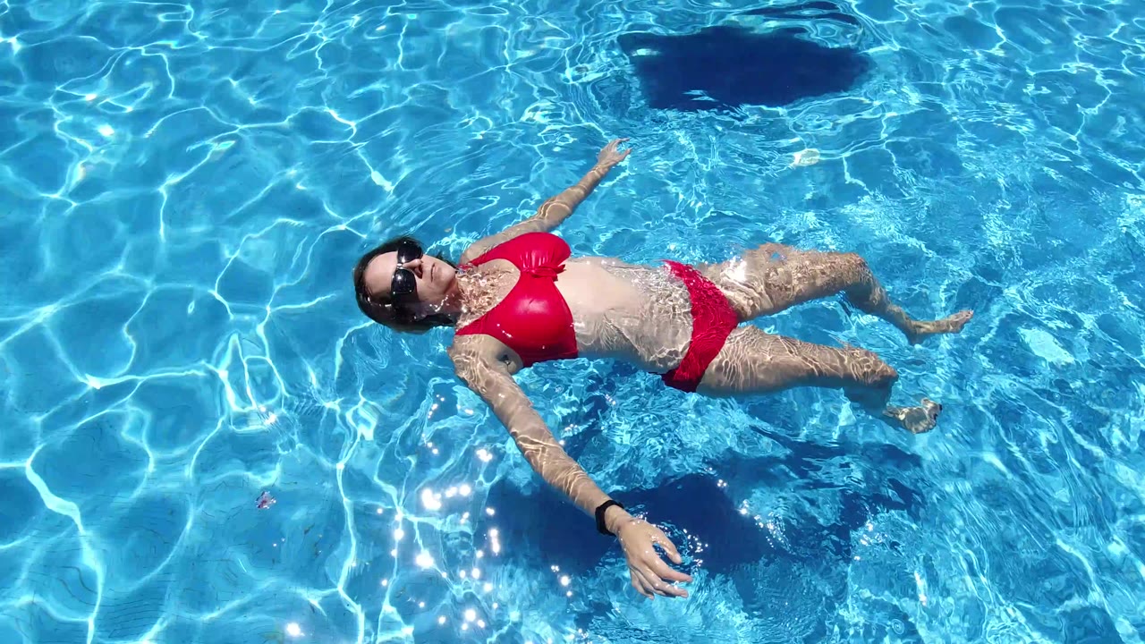 Overhead frame from Los Cabos, Mexico showing an adult woman in a red bikini and sunglasses floating on her back in clear blue pool water near a dark pool feature.
