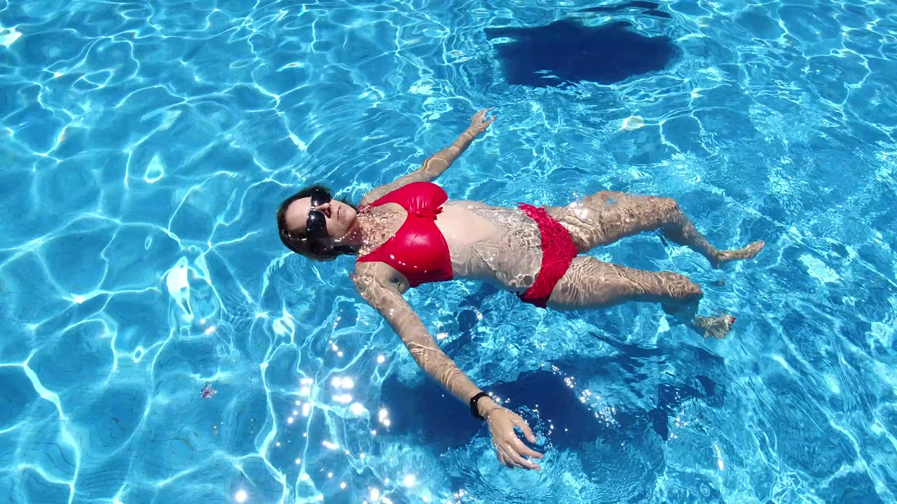 Overhead Los Cabos pool frame with the woman in a red bikini drifting on her back, one arm extended, and bright caustic light patterns across the blue water.