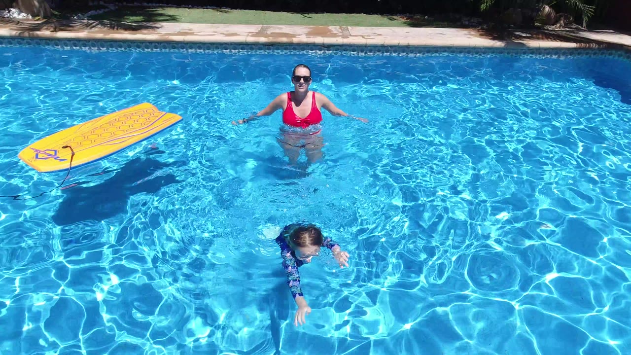 High angle view in Los Cabos, Mexico of a woman in a red swimsuit standing in a blue pool while a child swims below her and a yellow float board sits near the left edge.