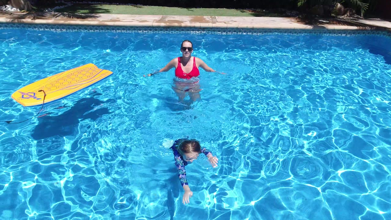 The Los Cabos pool remains framed from above as the woman faces camera near center, the child swims lower in the water, and the yellow float board drifts along the left side.