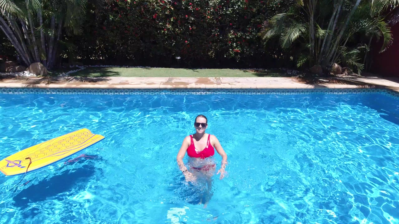 The woman remains centered in the Los Cabos swimming pool while the yellow float sits farther left on rippled blue water and the shaded hedge lines the background.