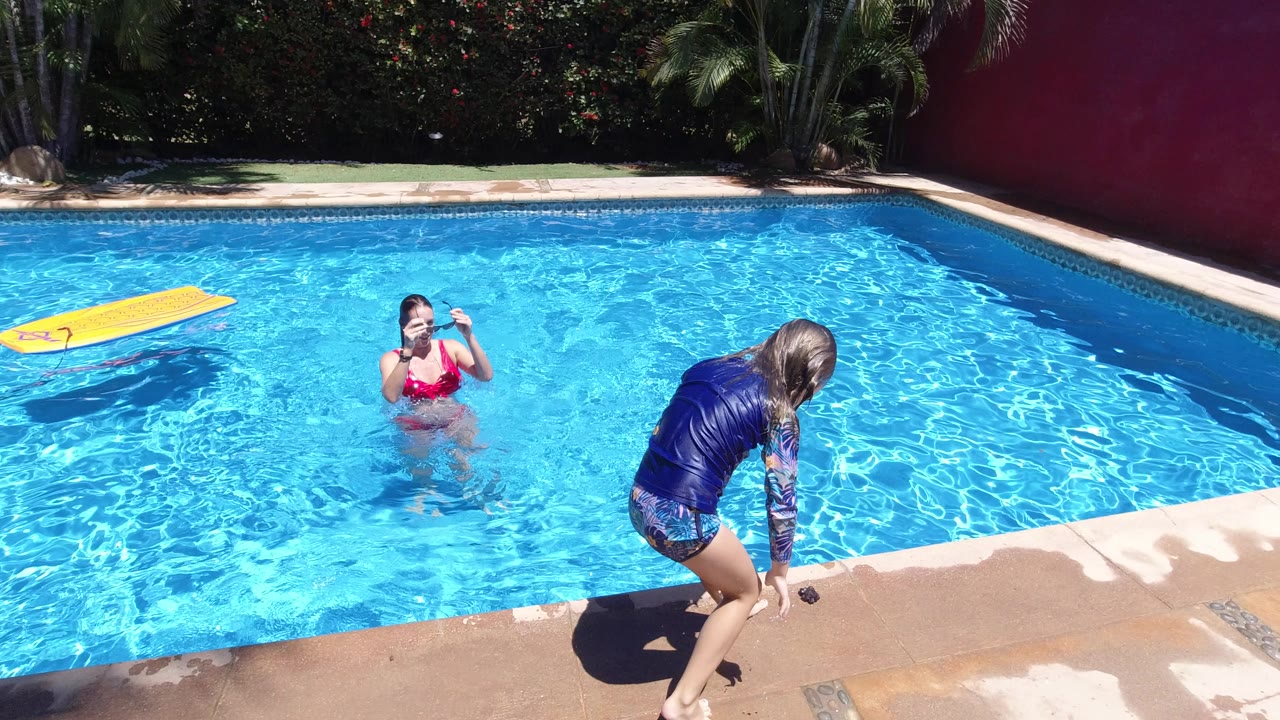 In Los Cabos, Mexico, a young girl in a blue rash guard stands on the wet pool deck while a woman in a red swimsuit faces her from the bright blue water.