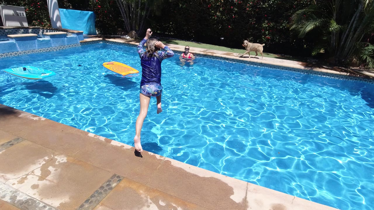 Child mid-jump toward a bright blue swimming pool with another child and dog visible.