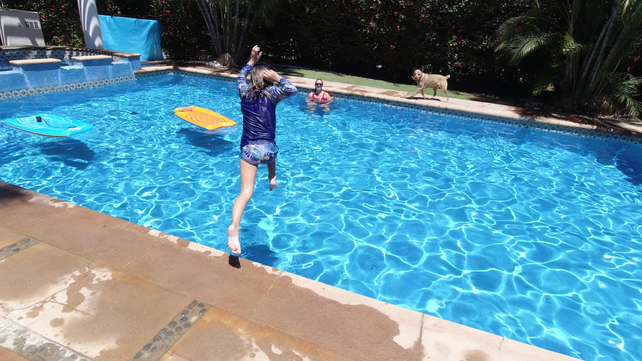 A child is airborne over the Los Cabos pool, with rippled blue water below, another child near the far edge, the dog on the coping, and two floats drifting nearby.