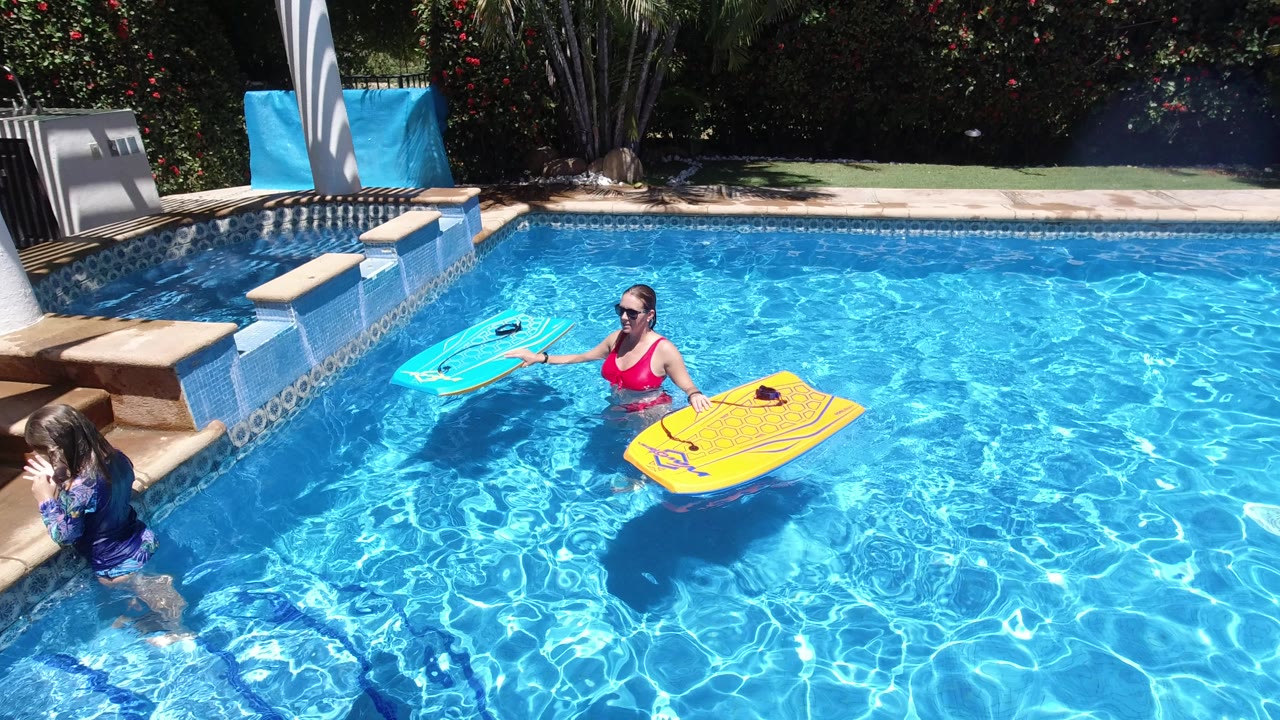 The Los Cabos pool scene continues with the woman centered in the water, the child at left by the pool wall, and two bright bodyboards floating beside her.