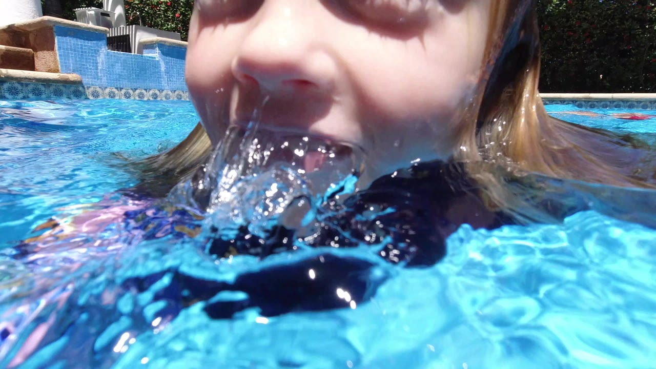 A close waterline view shows the blonde child smiling with water splashing at her mouth in a bright blue pool in Los Cabos, Mexico.