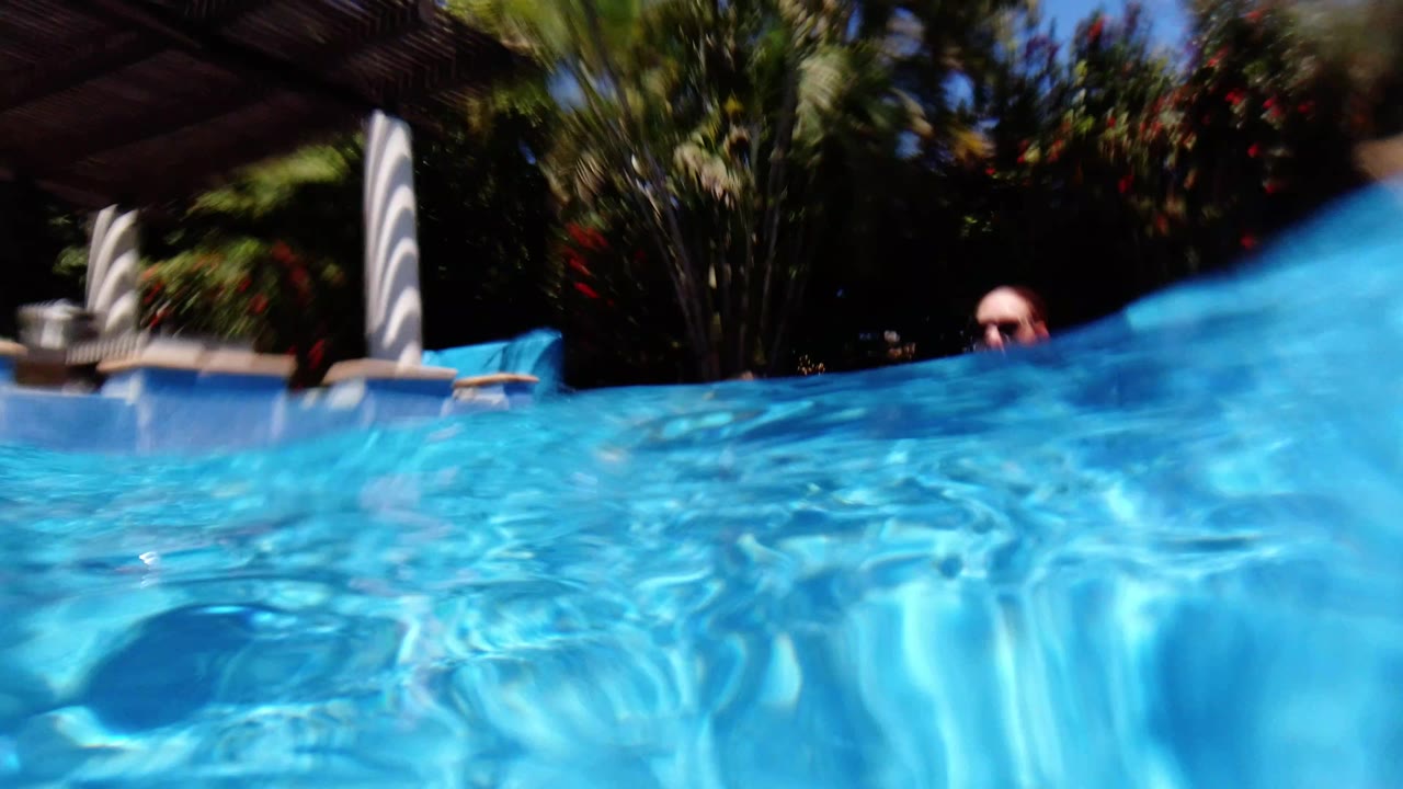 Partly submerged view across a Los Cabos pool with rippled blue water, shaded patio columns, greenery, and the top of a person barely visible near the waterline.