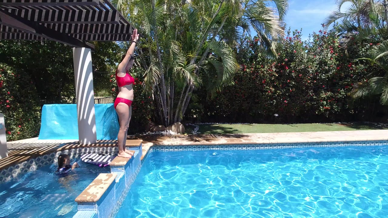 Woman in pink swimsuit standing on a pool ledge beside a swimmer.