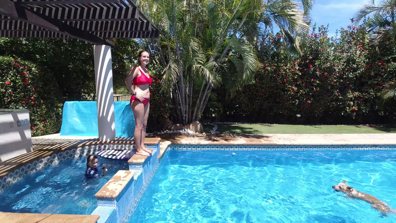 Woman in a red bikini stands on a pool ledge while a child and dog are in the water.