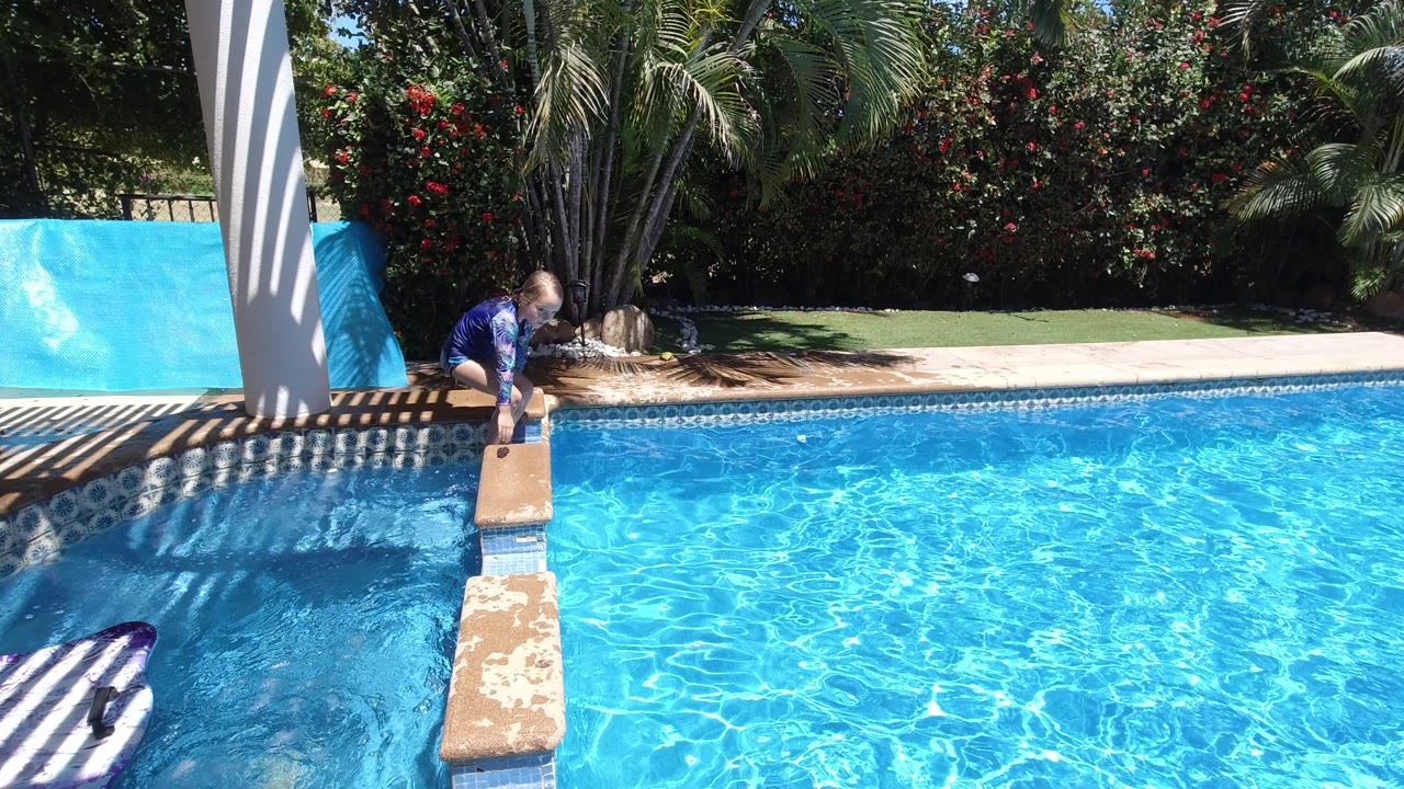 The child leans forward from the pool edge toward the small diving board while bright blue water fills most of the Los Cabos frame.