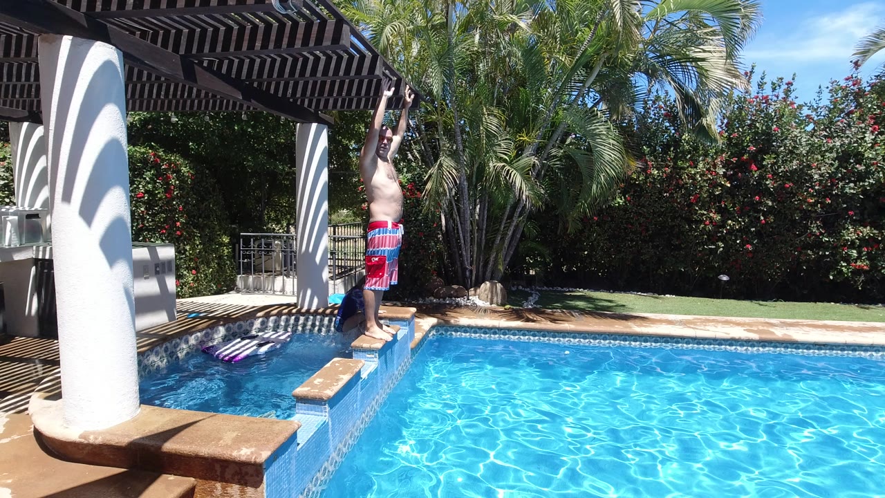 At 0:01 in Los Cabos, Mexico, the man holds his raised-arm stance on the tiled pool edge as the child stays close to the side of the pool.