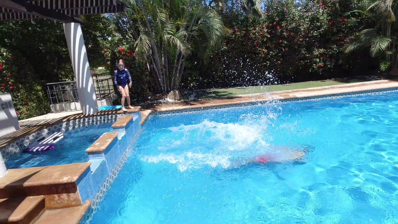 In Los Cabos, Mexico, a child stands near the pool edge as another child creates a large splash in the bright blue water, with striped shade, white columns, palms, and hedges behind them.