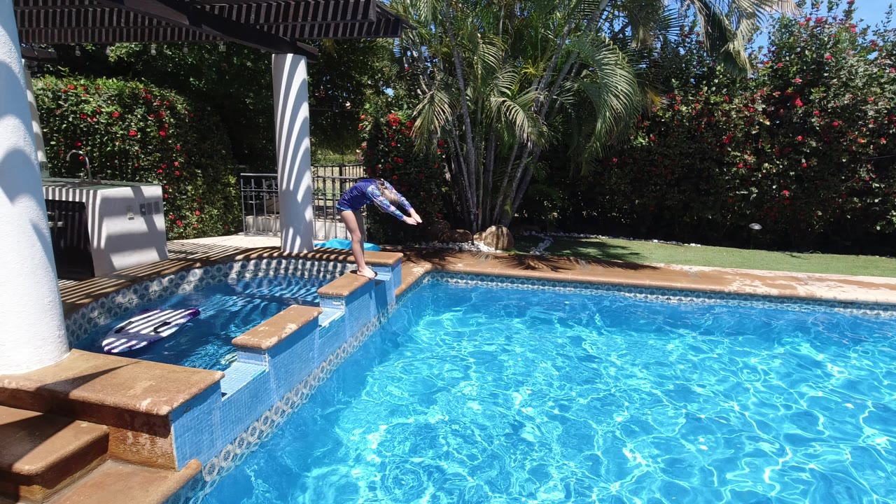 At 0:01, the girl holds her forward-leaning dive stance on the pool coping as sunlight patterns ripple across the blue water and the shaded patio sits at left.