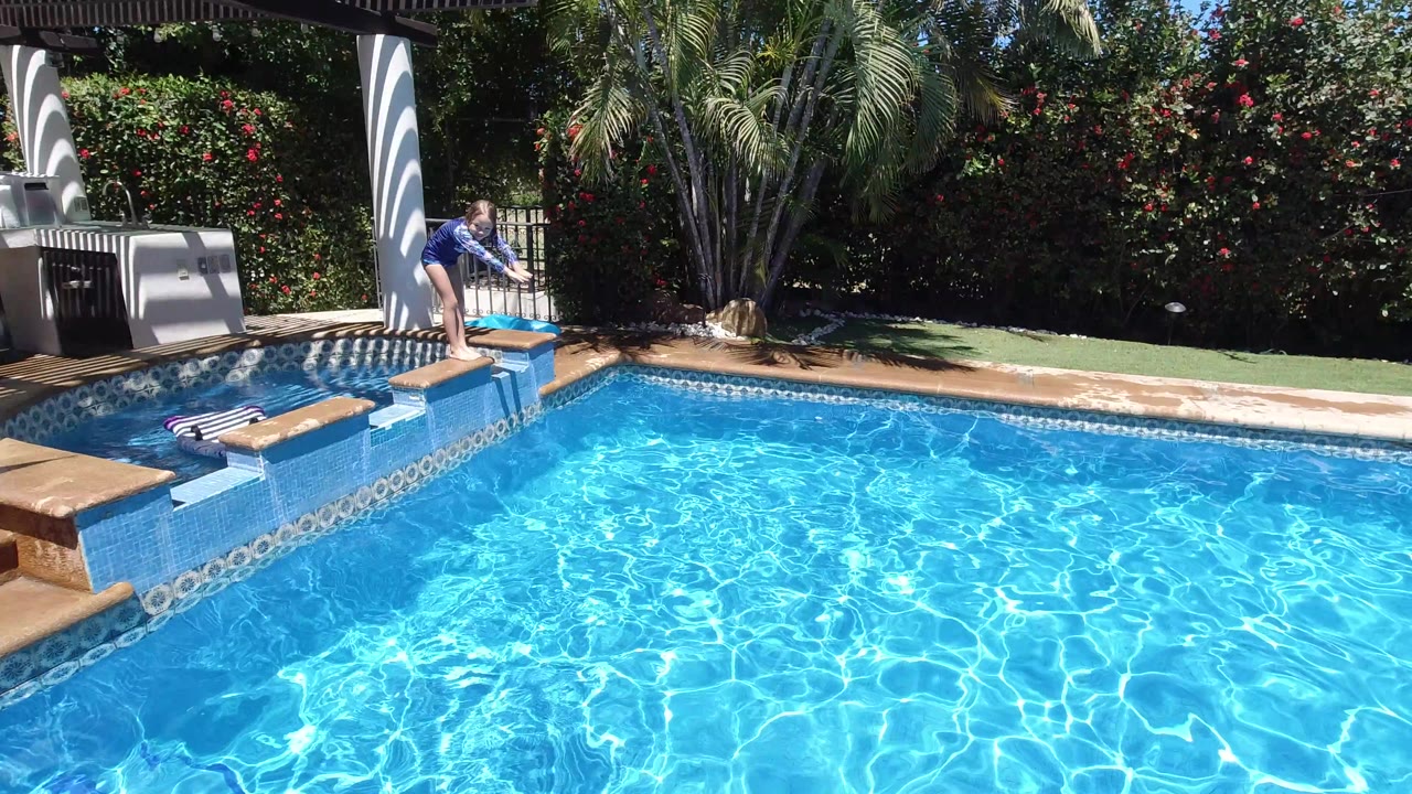 The child bends closer to the pool from the ledge in Los Cabos, with rippled turquoise water filling most of the frame and garden shrubs in the background.