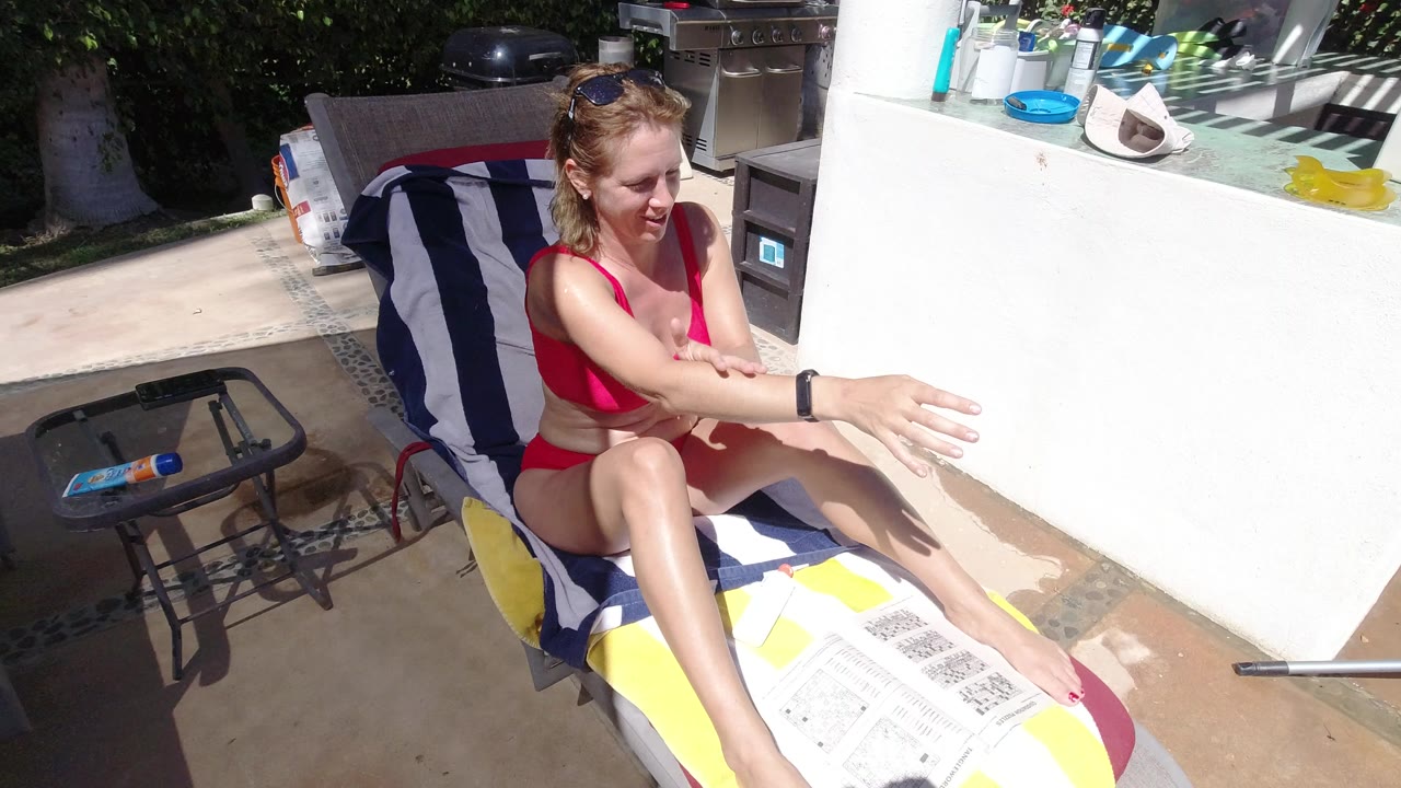 At 0:00 in Los Cabos, Mexico, a woman in a pink bikini sits on a striped lounge chair and rubs sunscreen along her extended forearm in bright patio sun.